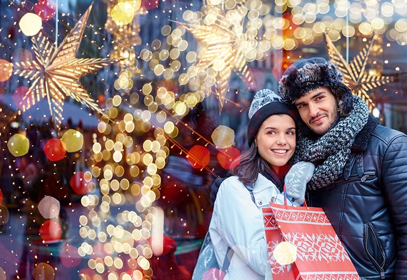 A happy couple stands together, smiling in front of a festive, illuminated store window adorned with holiday decorations.