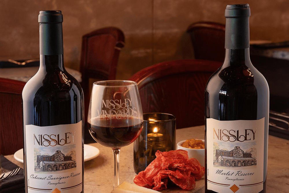 Two bottles of Nissley wine with a glass, accompanied by charcuterie, on a table with ambient lighting.