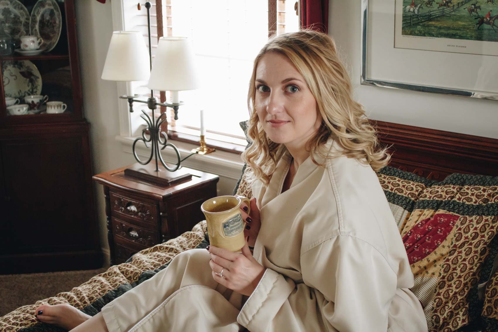 A woman in a beige robe sits on a bed, holding a mug and looking at the camera.