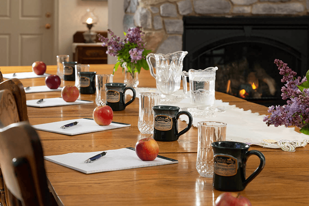 A wooden dining table set with pens, notepads, mugs, apples, and glass pitchers, with a fireplace in the background.