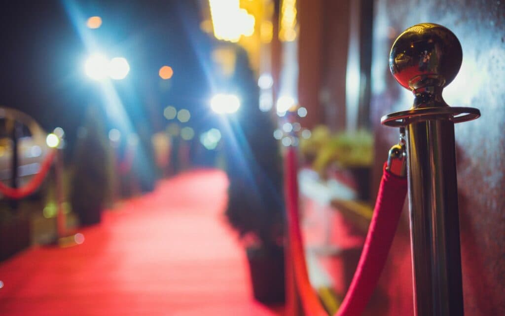 A polished stanchion with a red rope lines a dimly lit, glitzy red carpet leading into a venue.