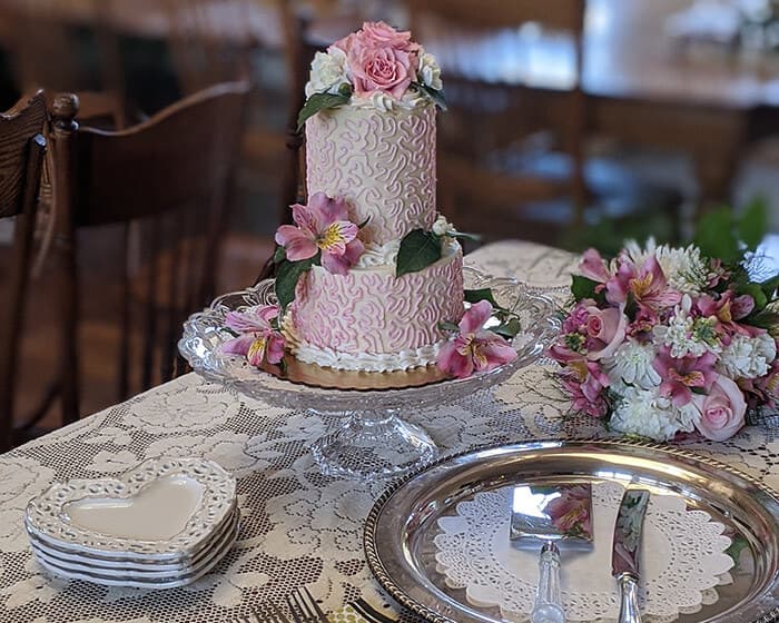 A beautifully decorated two-tiered cake adorned with flowers, displayed on a lace tablecloth alongside a floral bouquet and elegant tableware. A beautifully decorated two-tiered cake adorned with flowers, displayed on a lace tablecloth alongside a floral bouquet and elegant tableware.