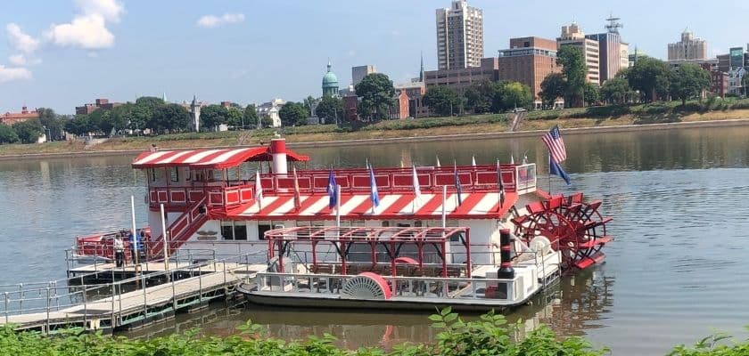 A red and white riverboat with a paddle wheel docked alongside a city waterfront.