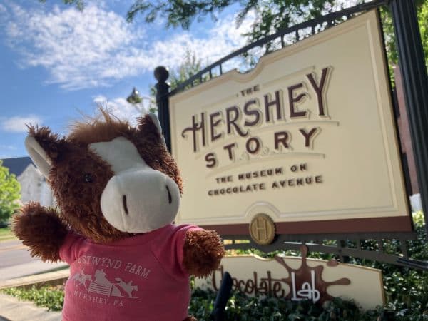 A plush horse in a pink shirt poses in front of the Hershey Story museum sign.
