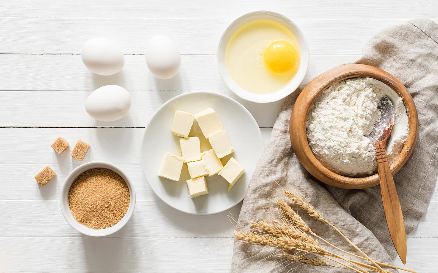 Ingredients for baking: eggs, butter, flour, sugar, and wheat stalks on a white surface.