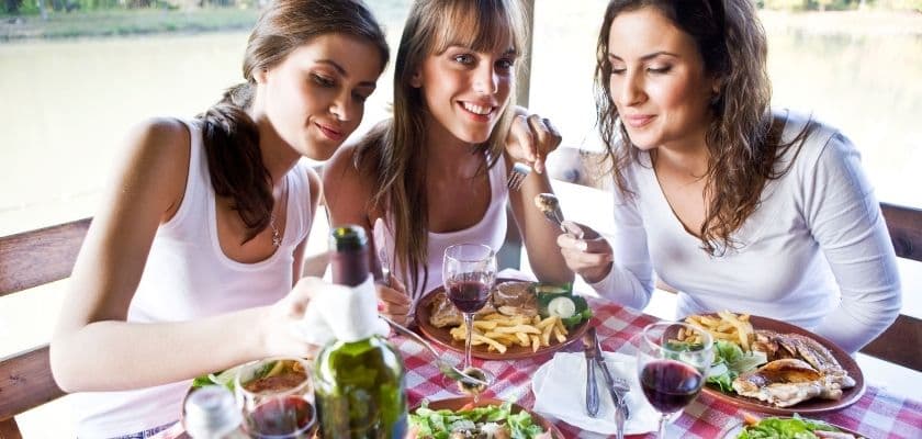 Three women enjoying a meal together outdoors by a lake.