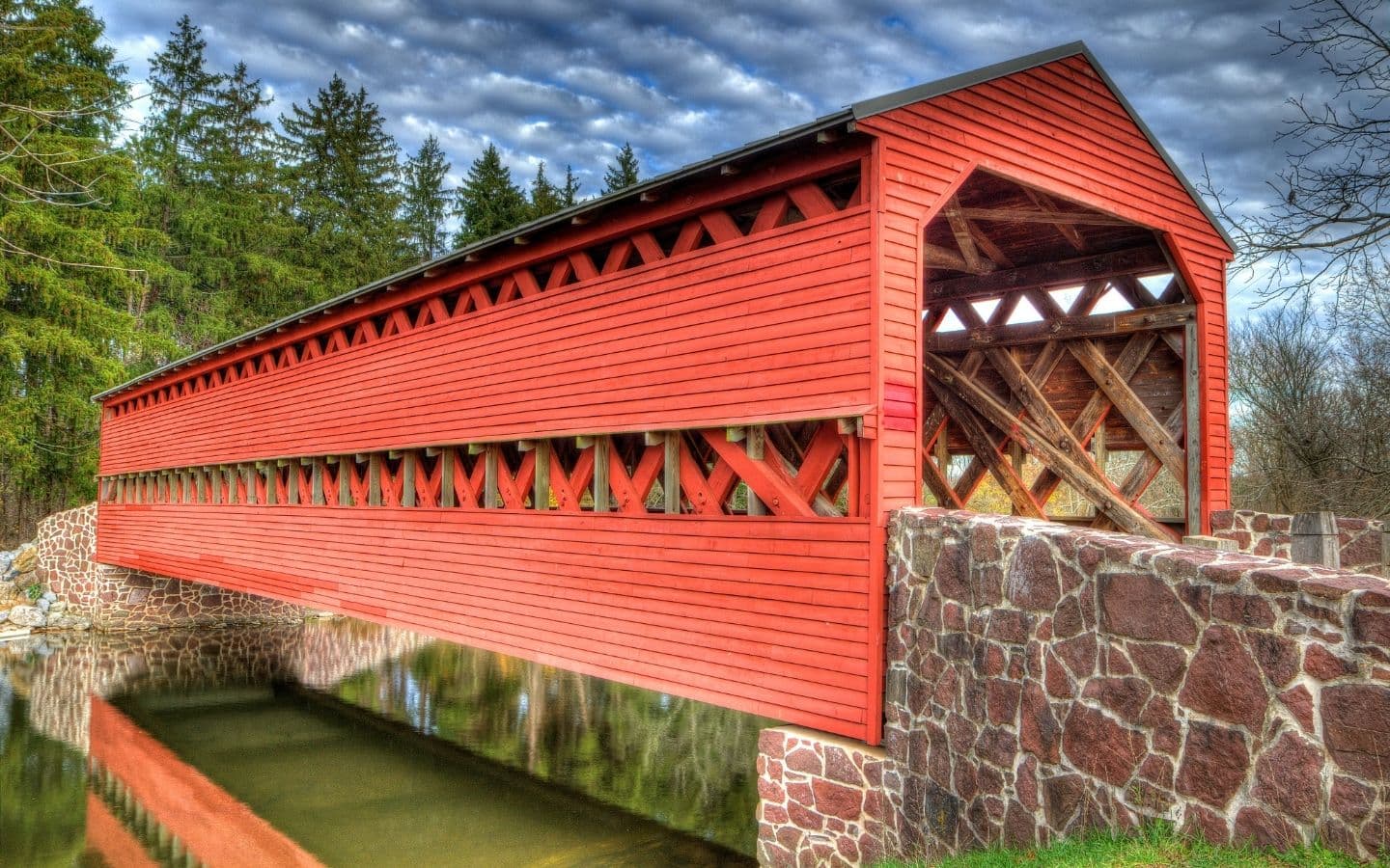 A red covered bridge spans over a calm body of water with trees in the background.