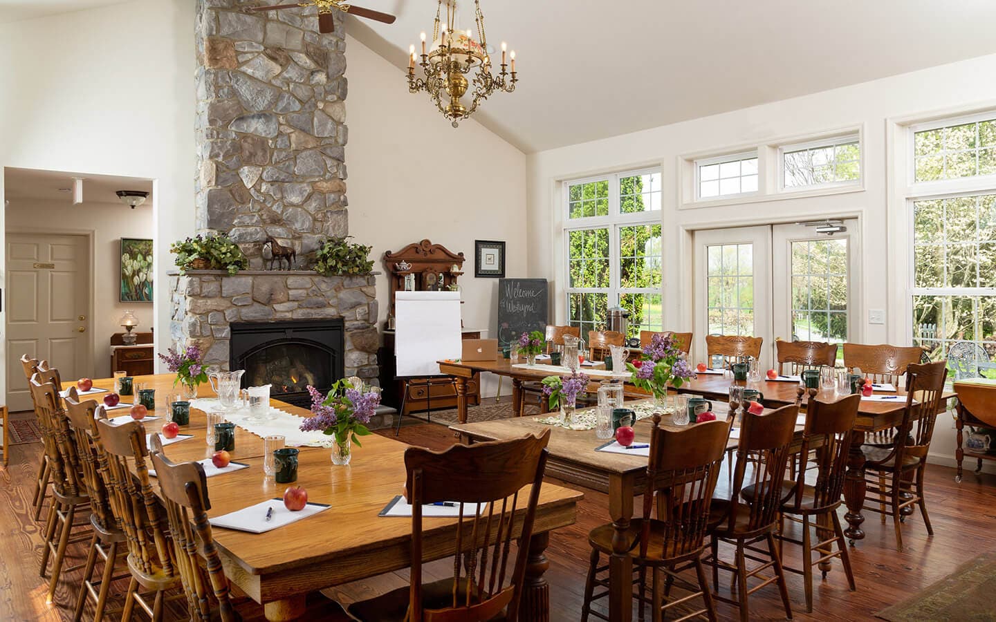 A spacious meeting room featuring a long wooden table, stone fireplace, and arrangements of flowers and beverages.
