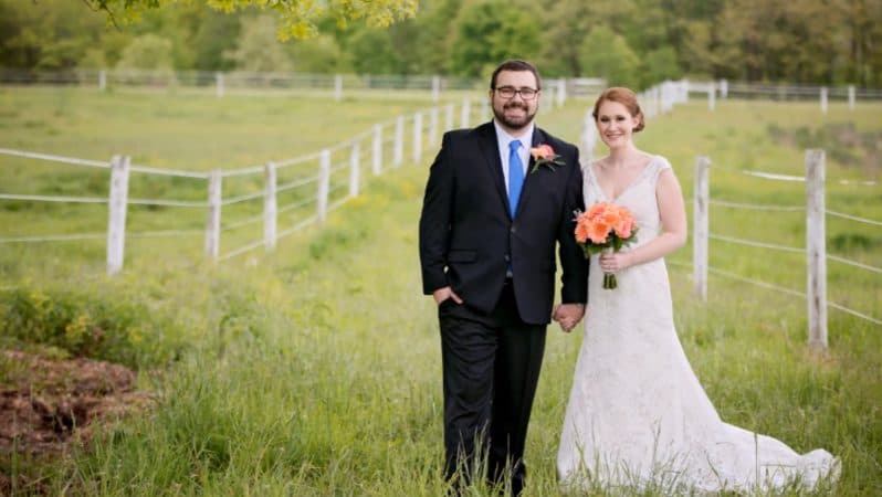 A smiling couple stands in a grassy field, holding hands and dressed in wedding attire.