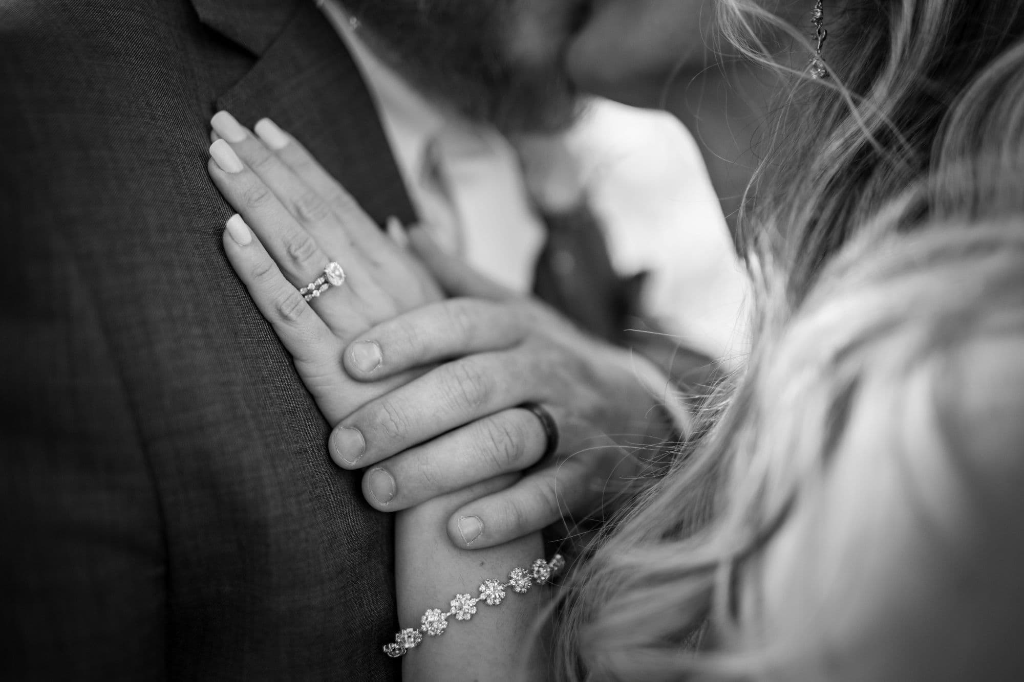 A close-up of two hands intertwined, showcasing wedding rings and a bracelet, with a couple embracing in the background.