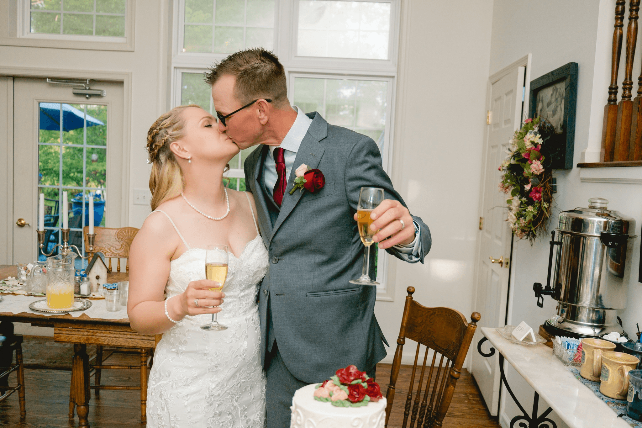 A newlywed couple shares a kiss while holding champagne glasses in a decorated interior.