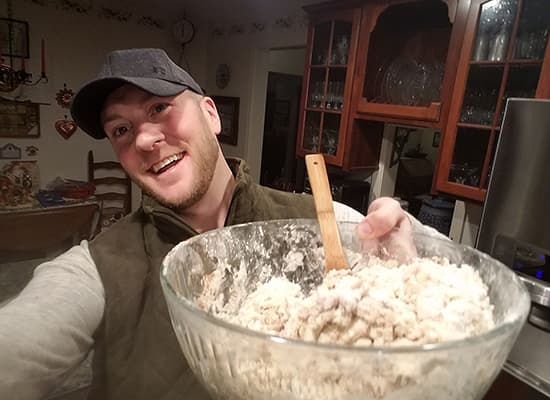 A man smiles while holding a large glass bowl filled with dough in a cozy kitchen.
