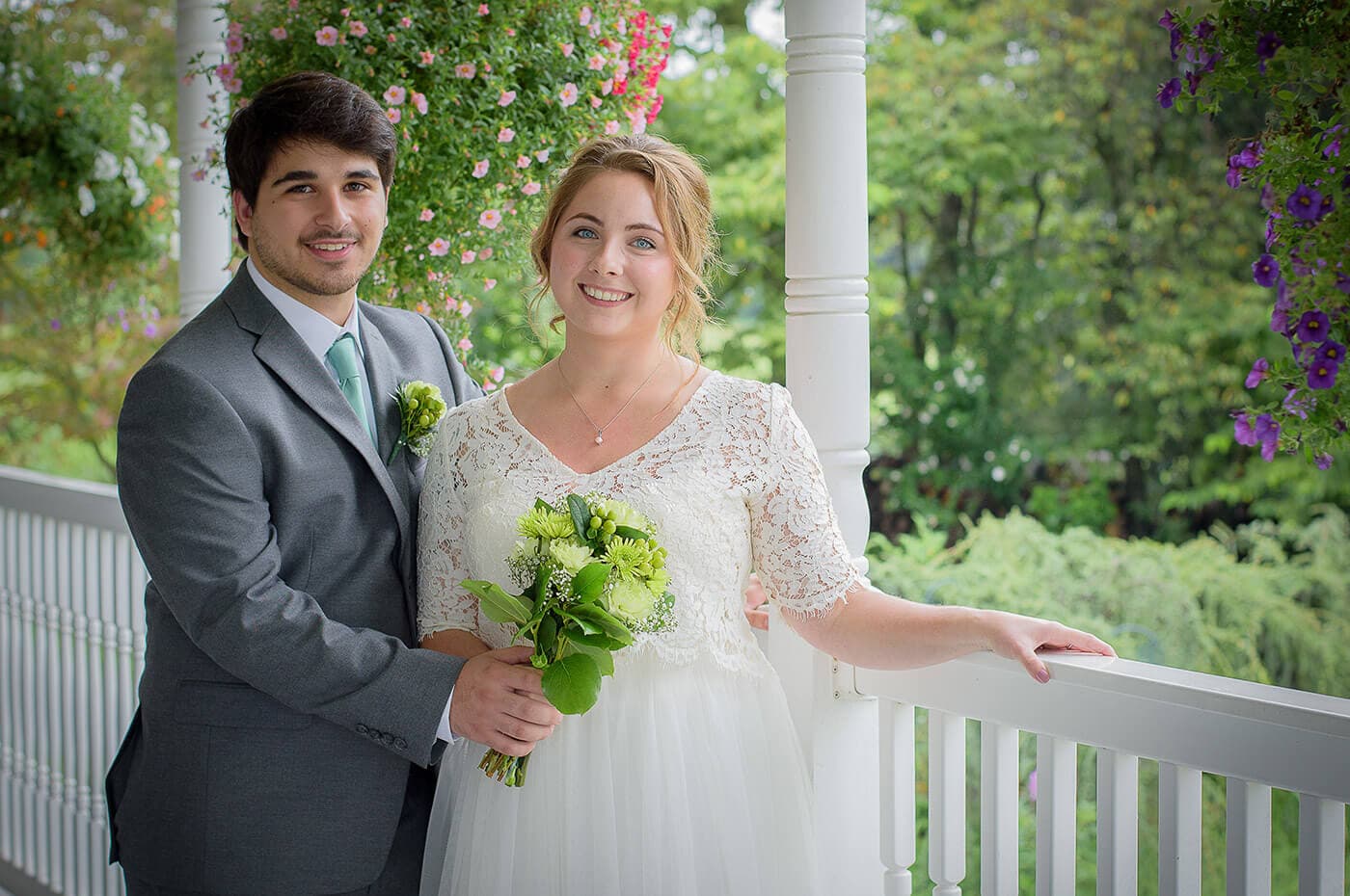 A couple in formal attire stands together on a porch, smiling while holding a bouquet and surrounded by hanging flowers. A couple in formal attire stands together on a porch, smiling while holding a bouquet and surrounded by hanging flowers.