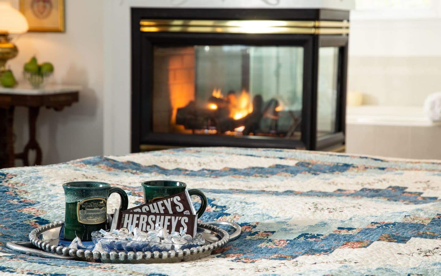 A cozy setting featuring two mugs, a Hershey's chocolate bar, and silver-wrapped candies on a quilted table, with a fireplace in the background.