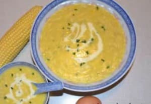 A bowl and a small bowl of creamy corn soup, garnished with herbs, alongside a corn cob and an egg.