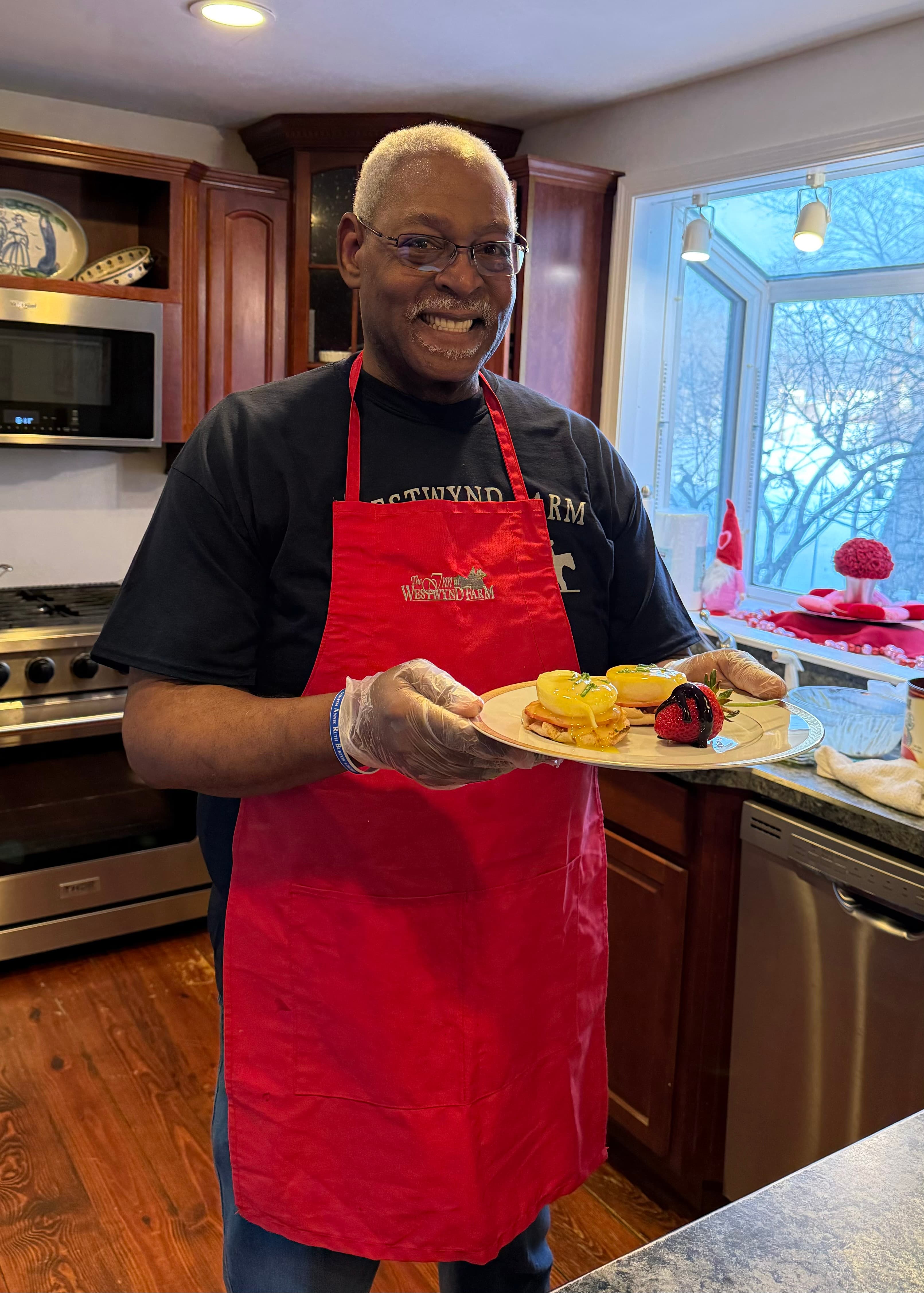 A smiling man in a red apron holds a plate of colorful food in a modern kitchen.