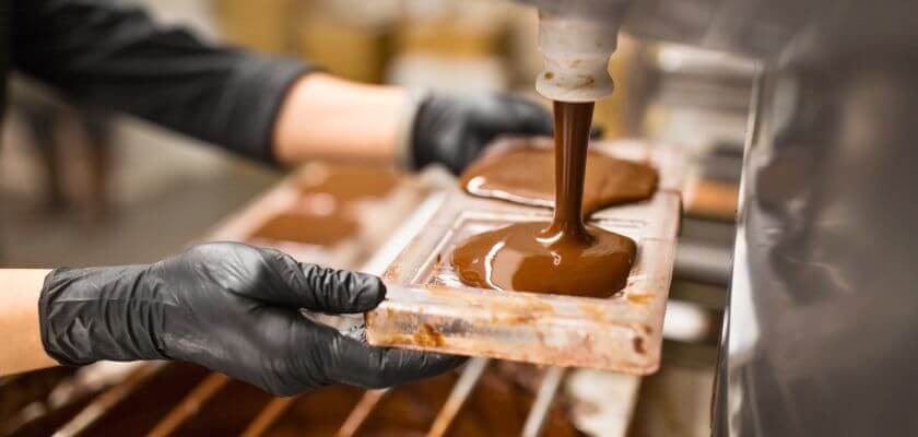 A person in black gloves pours melted chocolate into mold trays.