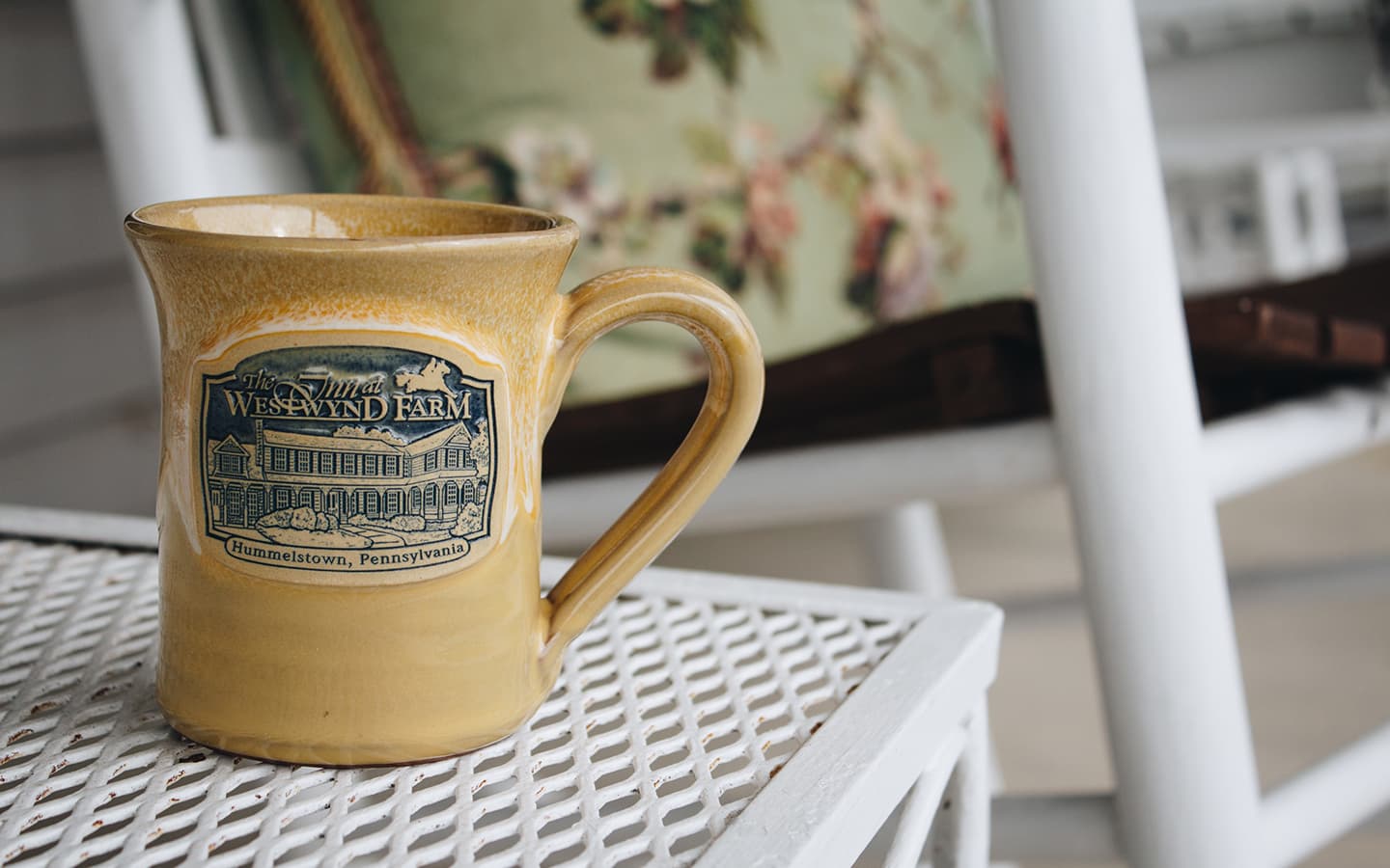 A ceramic mug featuring a design of the Inn at Westwynd Farm rests on a woven table.