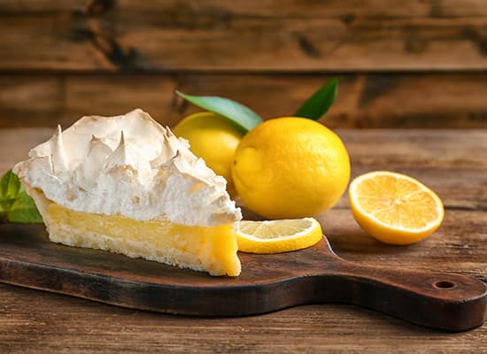 A slice of lemon meringue pie on a wooden board, accompanied by whole and sliced lemons.