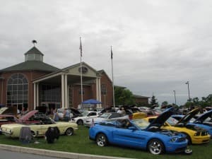 A variety of classic cars are displayed outside a large building under overcast skies.