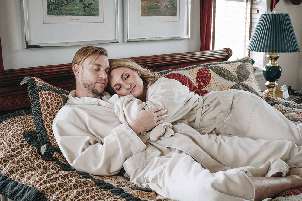 A couple in beige robes cuddles on a bed, surrounded by decorative pillows and soft lighting.