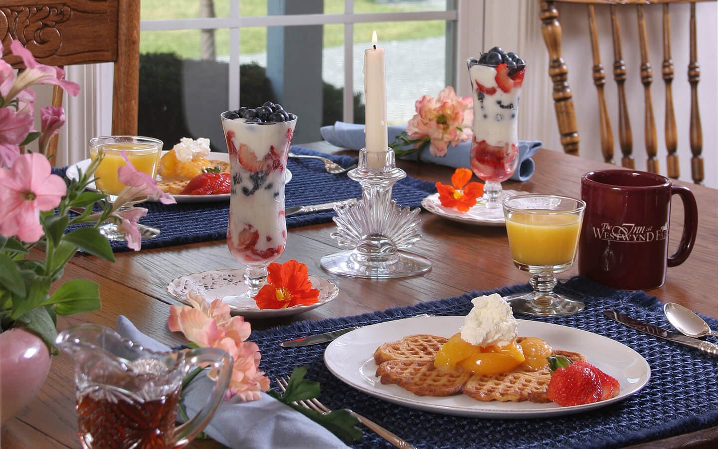 A beautifully set breakfast table featuring fruit parfaits, waffles with whipped cream and fruit, alongside drinks and flowers.