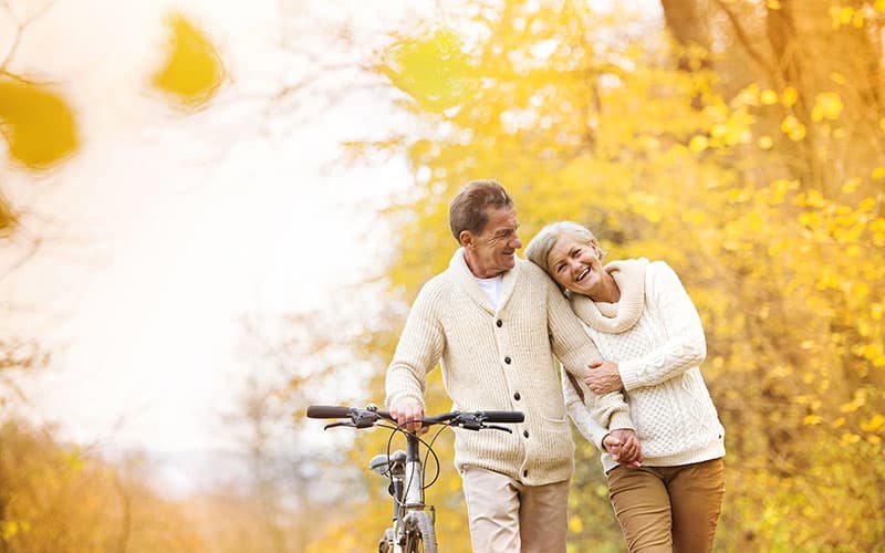 An elderly couple strolls hand-in-hand along a tree-lined path adorned with autumn leaves.