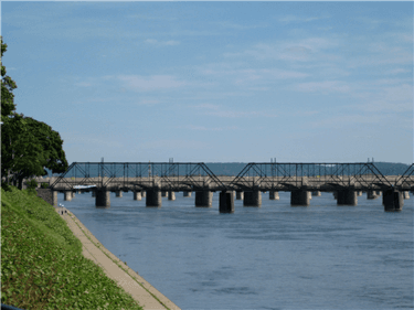 A metal bridge spans a calm river under a clear blue sky.