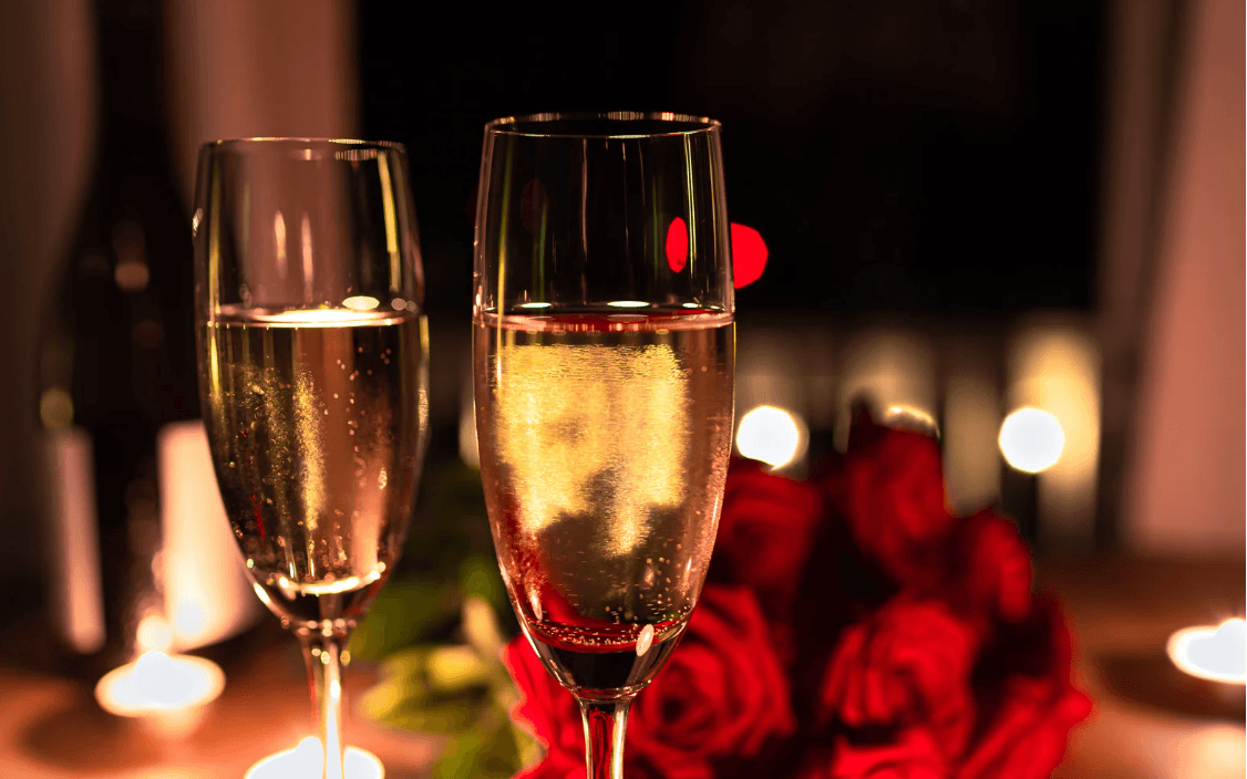 Two flutes of sparkling champagne beside a bouquet of red roses and soft candlelight.