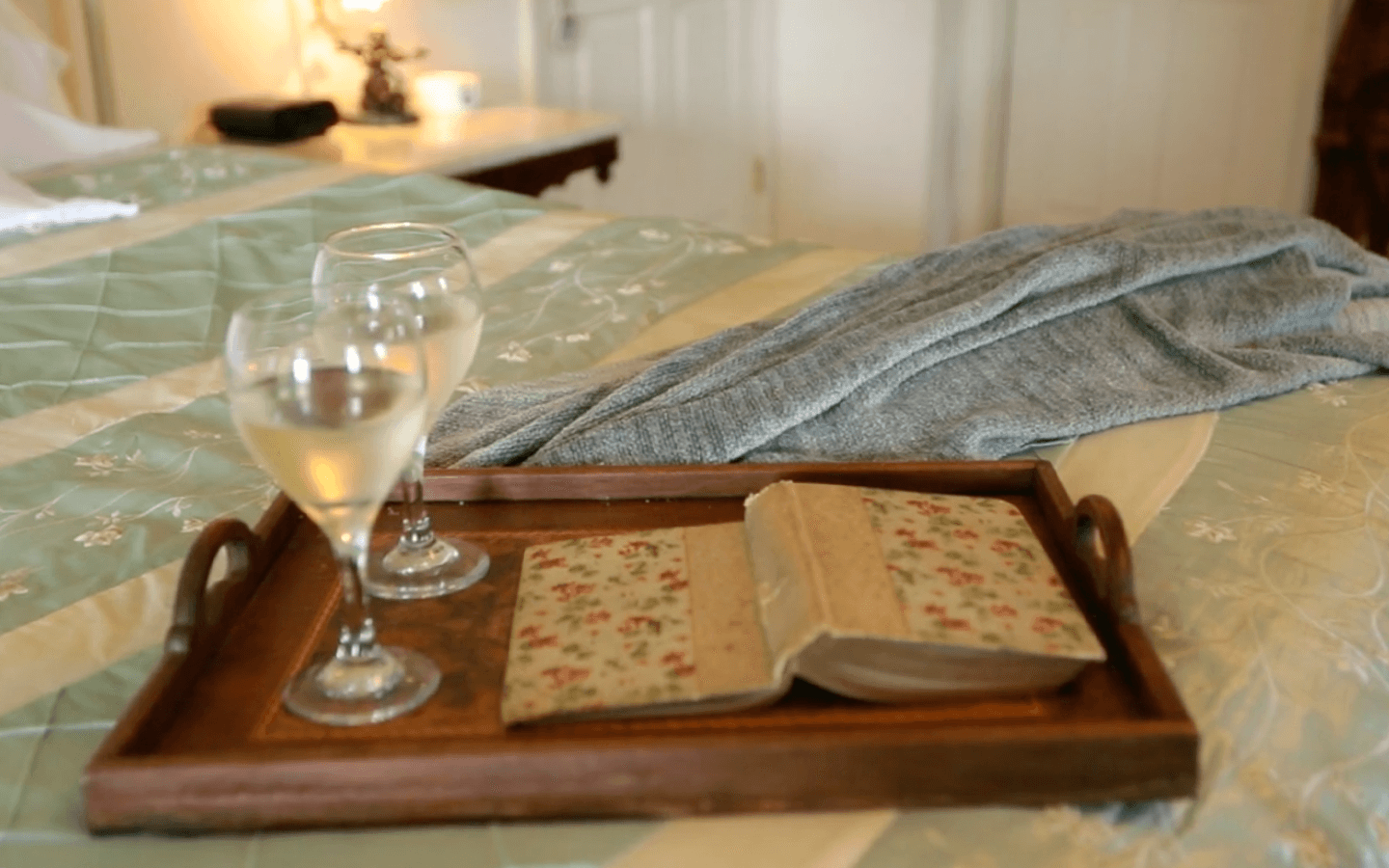 A wooden tray holds two glasses of white wine and an open floral-covered book on a neatly made bed.