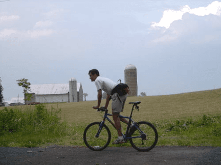 A person leans on a bicycle in front of a rural landscape, featuring a field and two silos.