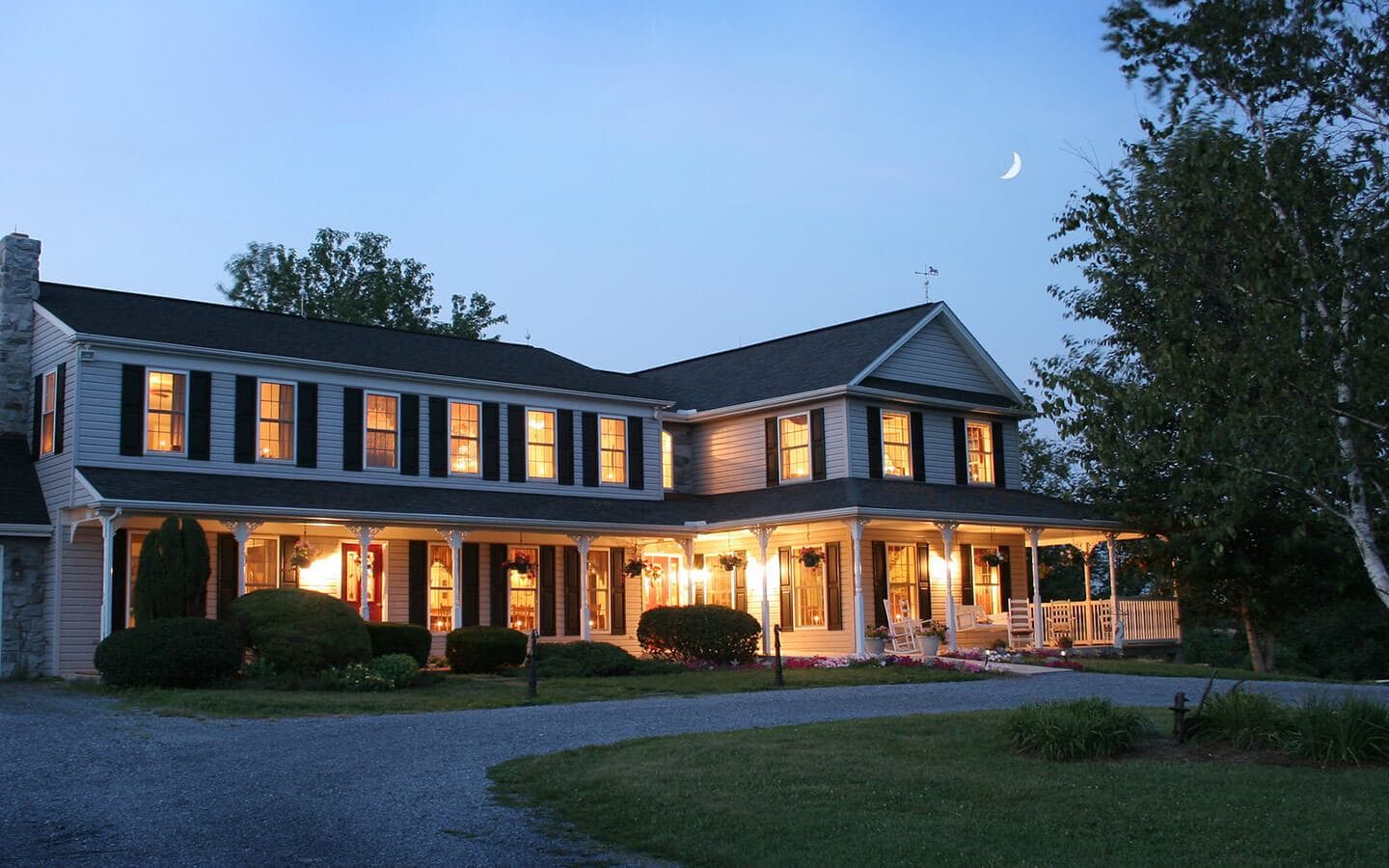 A charming two-story house illuminated at dusk, surrounded by lush greenery and a crescent moon overhead.