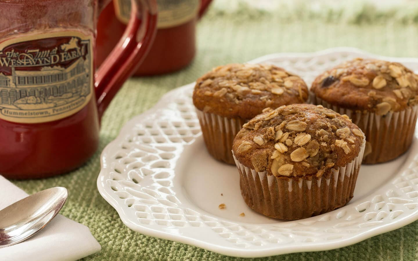 Three oatmeal muffins on an ornate white plate next to a red mug on a green tablecloth.