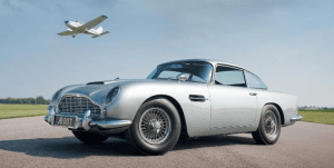 A classic silver car parked on an airstrip with a small plane flying overhead.