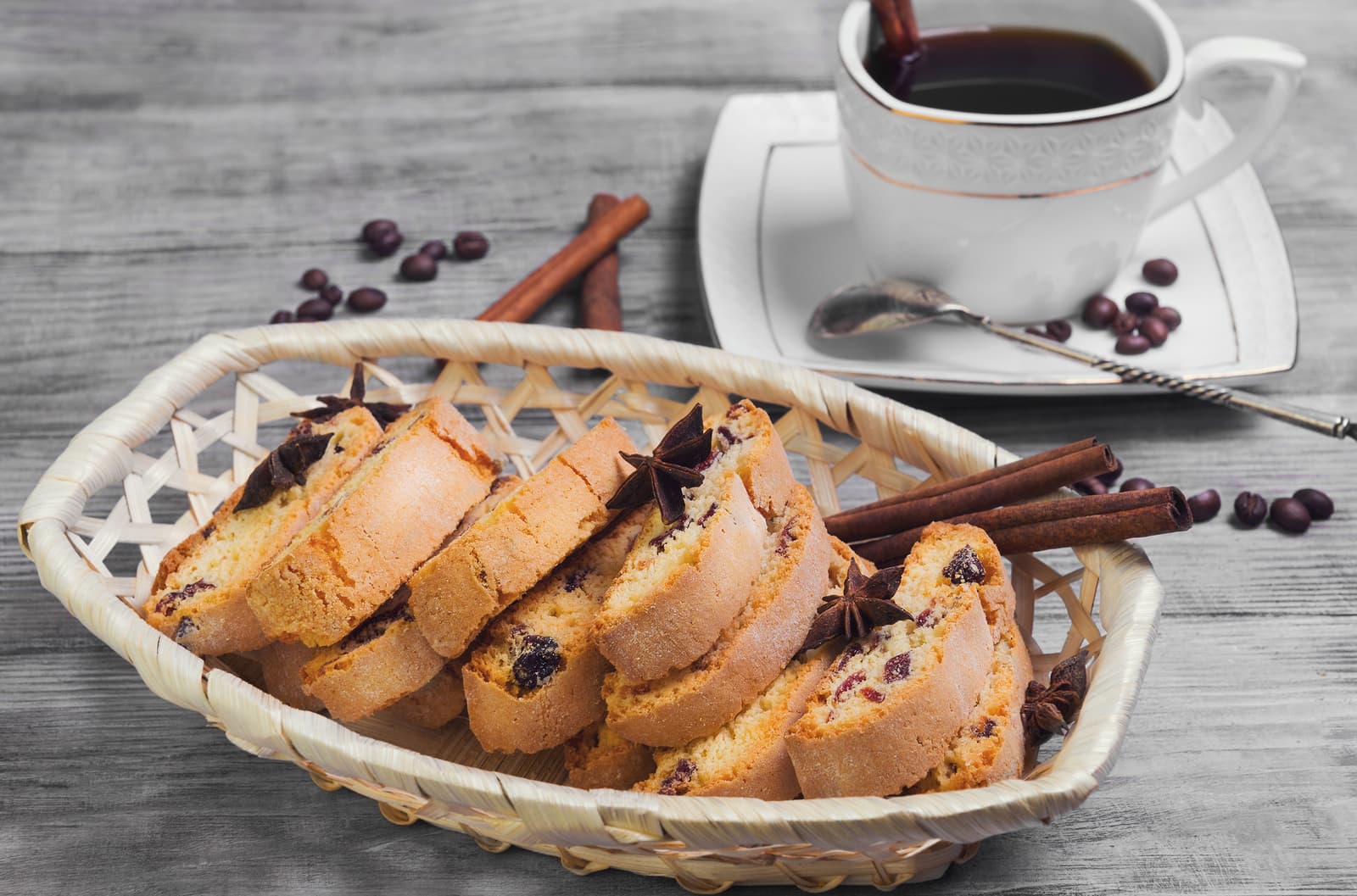 A basket of sliced cookies beside a cup of coffee and cinnamon sticks.