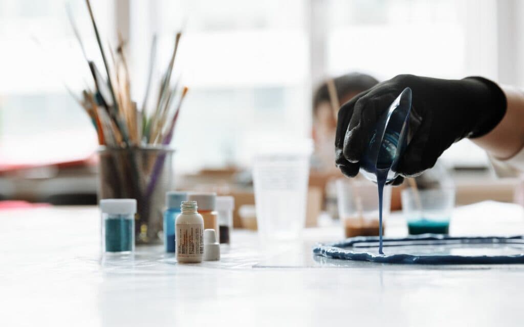 A gloved hand pours blue paint onto a table surrounded by paint containers and brushes.
