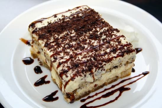 A slice of dessert with layers of cream, chocolate, and a drizzle of chocolate sauce on a white plate.