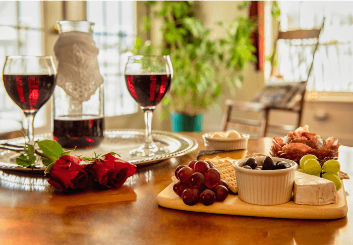 A beautifully arranged table with two glasses of red wine, a carafe, assorted cheeses, olives, and grapes, accompanied by red roses.