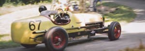 A yellow vintage racing car with the number 67 speeds around a curve on a rural road.