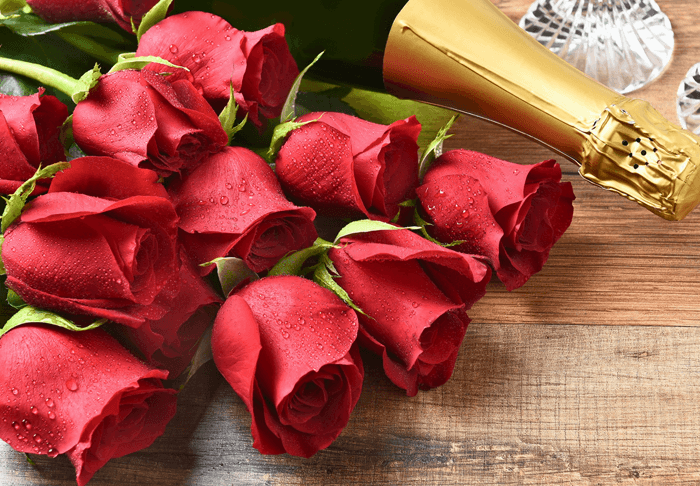 A cluster of red roses with droplets, resting near a champagne bottle and crystal glassware on wooden surfaces.