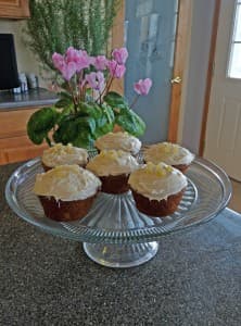 A glass cake stand holds six decorated cupcakes next to a potted plant with pink flowers.