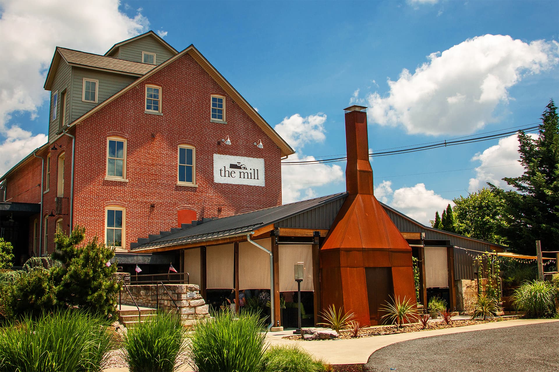 A rustic brick building with a sign reading "the mill," surrounded by greenery and under a blue sky.