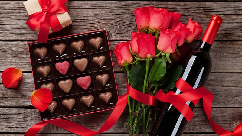 A gift arrangement featuring a box of heart-shaped chocolates, a bouquet of red roses, and a bottle of red wine on a wooden surface.
