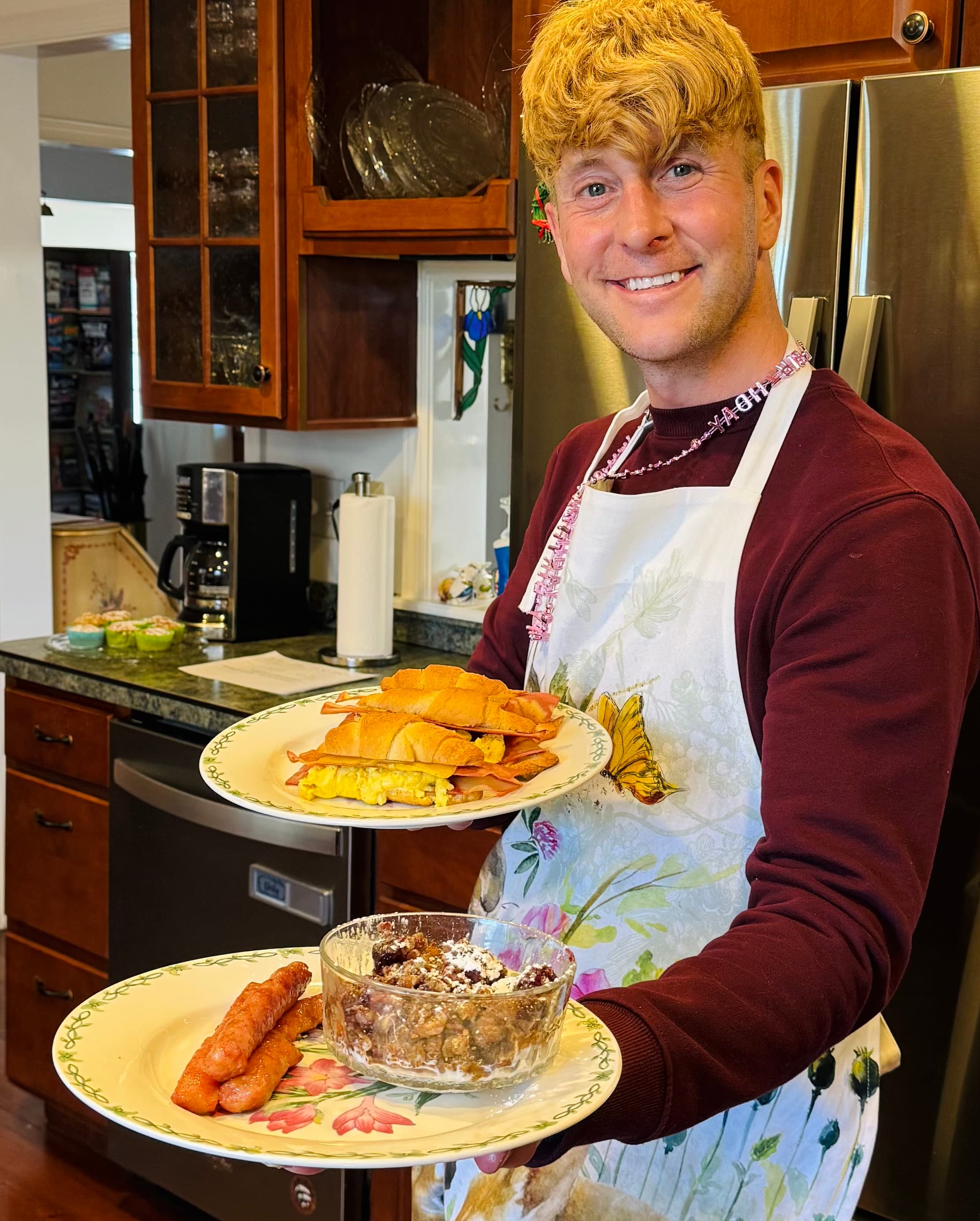 A smiling person wearing an apron holds two platters of food in a kitchen.