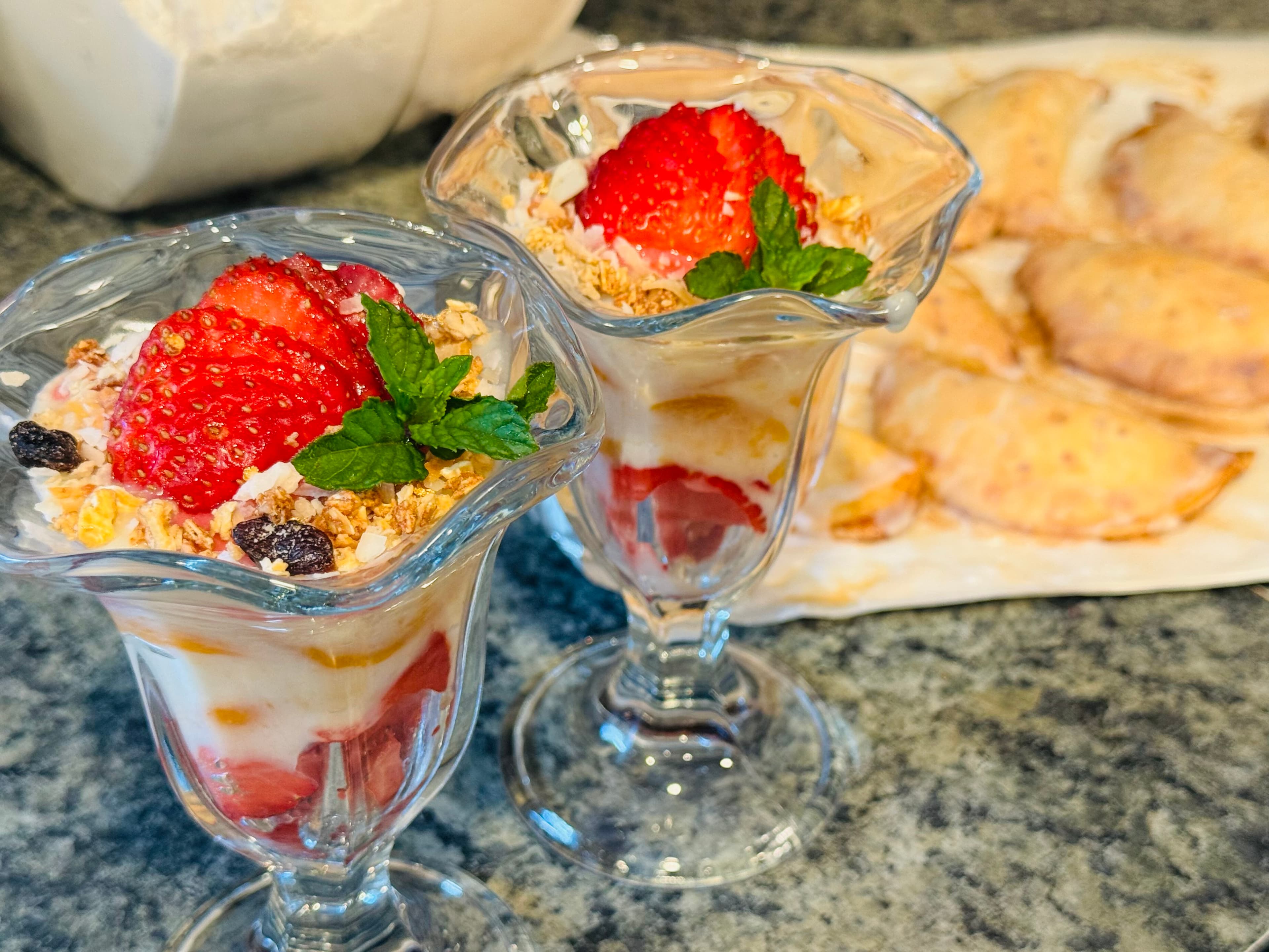 Two glasses of layered parfait with strawberries, granola, and mint, next to a plate of baked pastries.