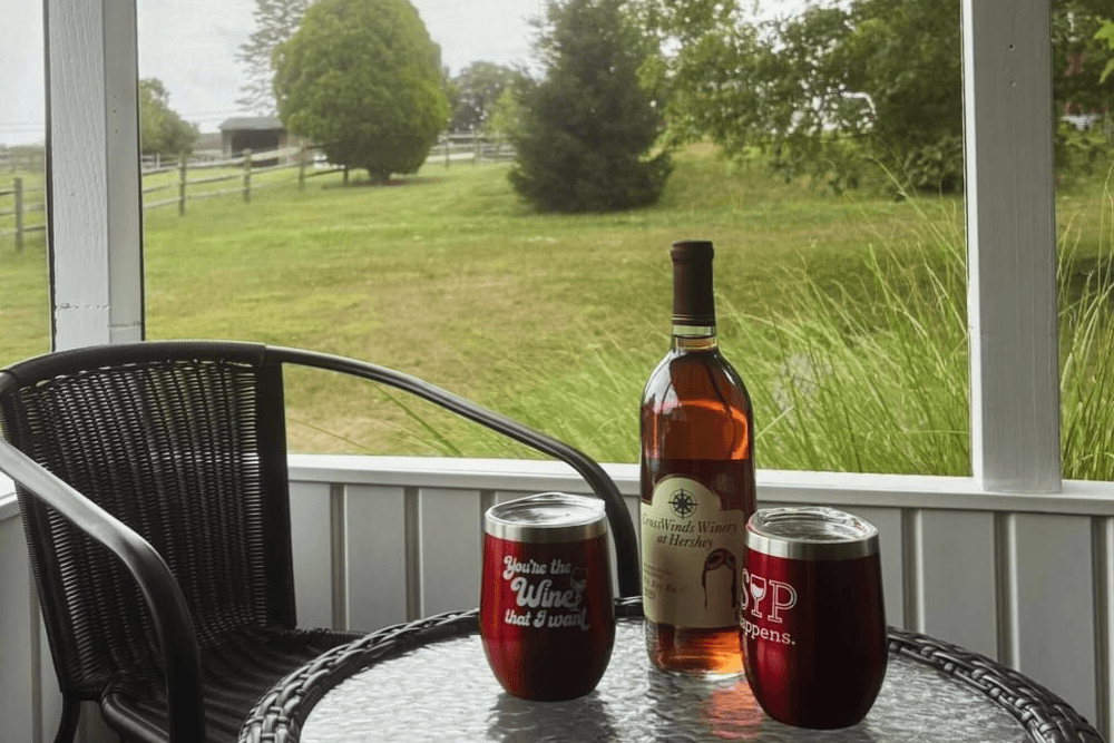 A bottle of wine sits on a glass table beside two insulated cups, with a green landscape in the background.