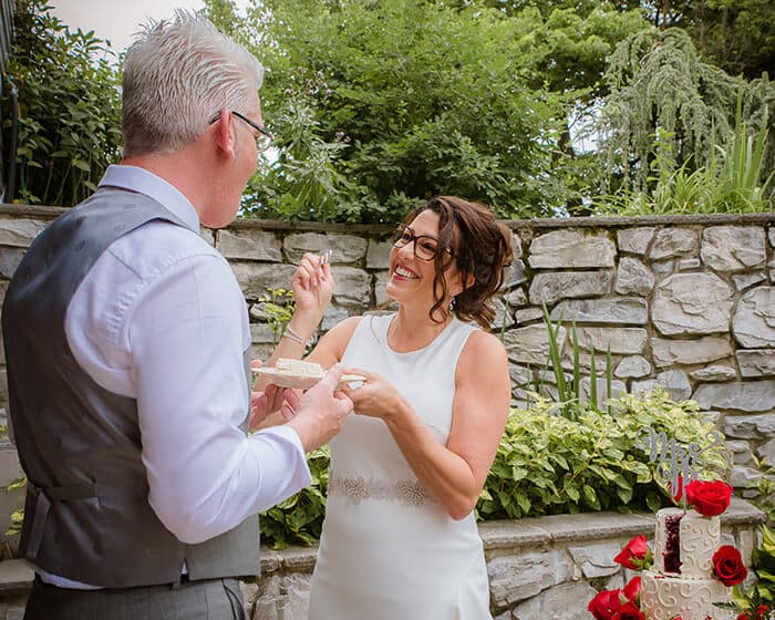 A smiling bride playfully offers cake to her groom in a lush outdoor setting.