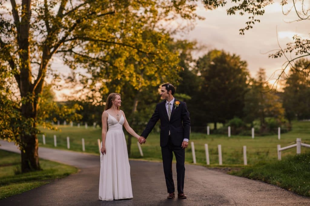 A bride and groom walk hand in hand along a tree-lined path during sunset.