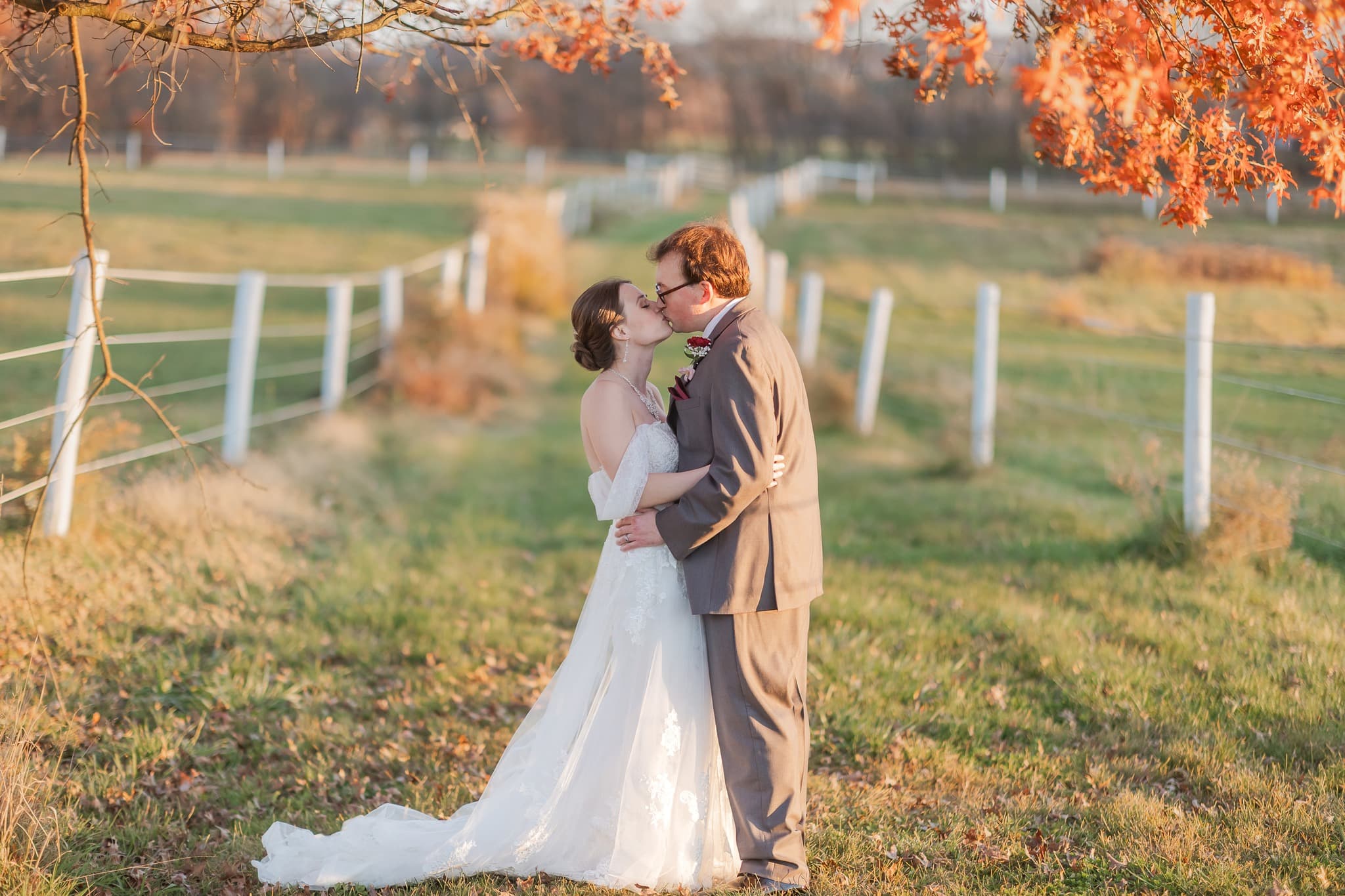 A bride and groom share a kiss in a picturesque outdoor setting with autumn foliage.