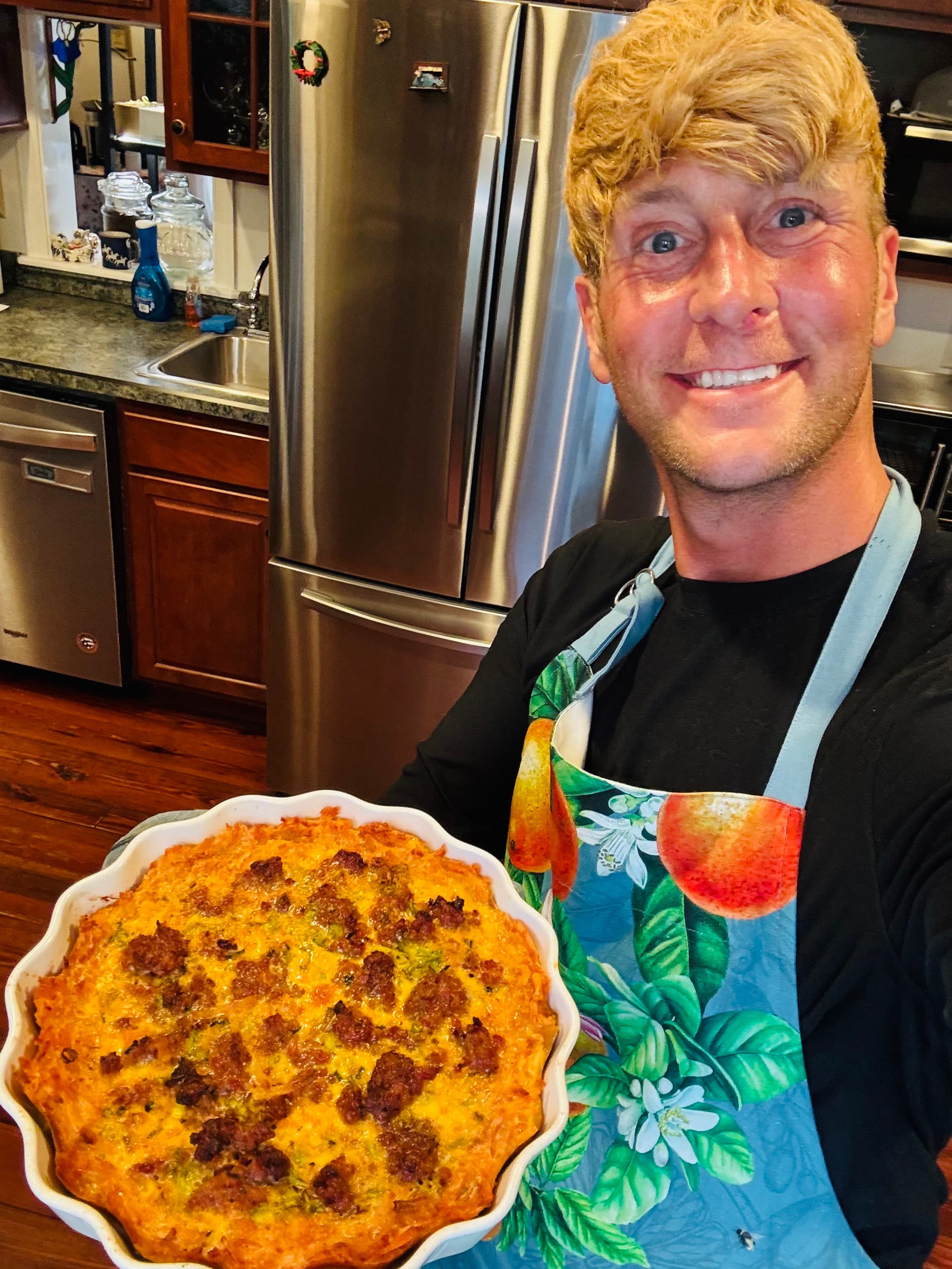 A smiling person in a colorful apron holds a freshly baked casserole in a kitchen.